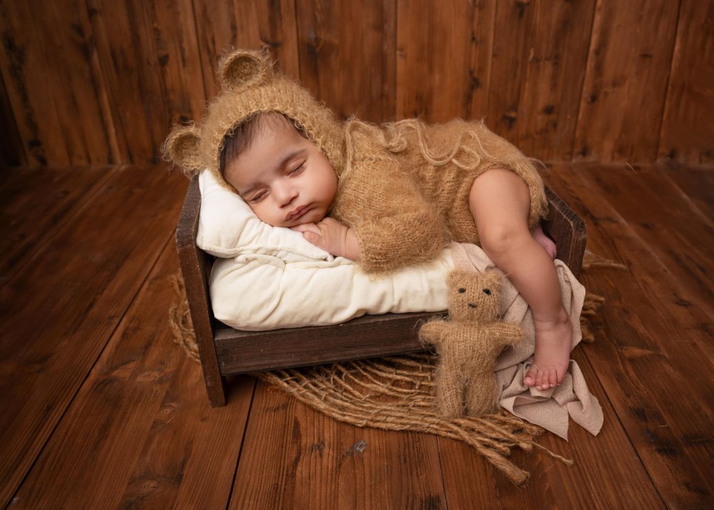 Charming photo of a newborn baby sleeping in a teddy bear outfit, set in a cozy wooden setting in Moga, India.