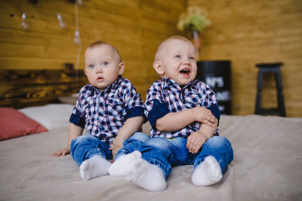 Cute twin boys in plaid shirts smiling and sitting on a bed indoors, capturing joy and sibling bond.