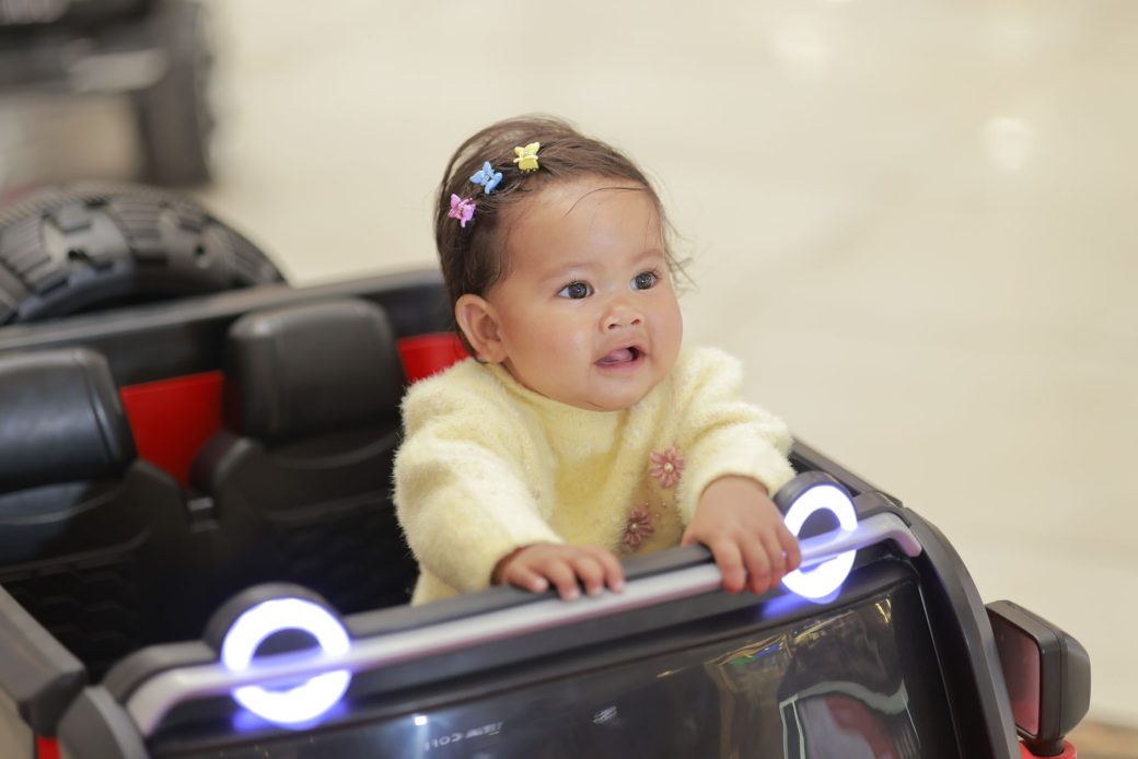 Cute baby in a toy car indoors, wearing colorful clips and a yellow sweater.