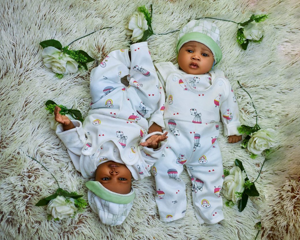 Cute twin babies in matching pajamas lying on a soft rug surrounded by white flowers.