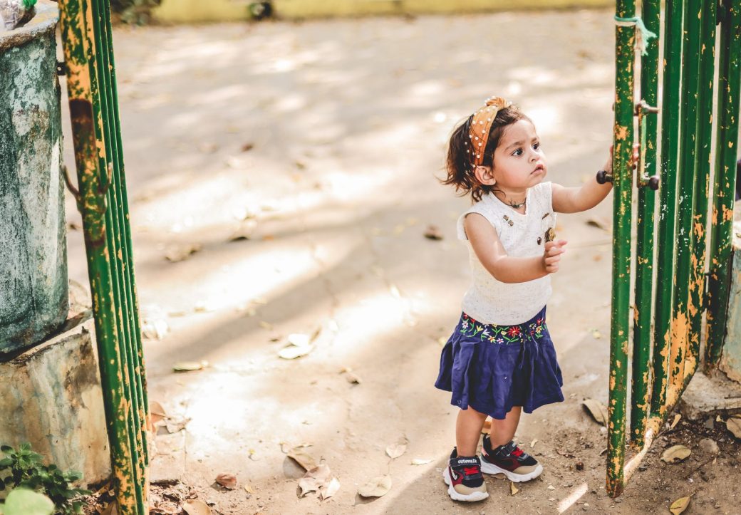 A young girl in a cute outfit is opening a vintage green gate in a sunlit outdoor setting.
