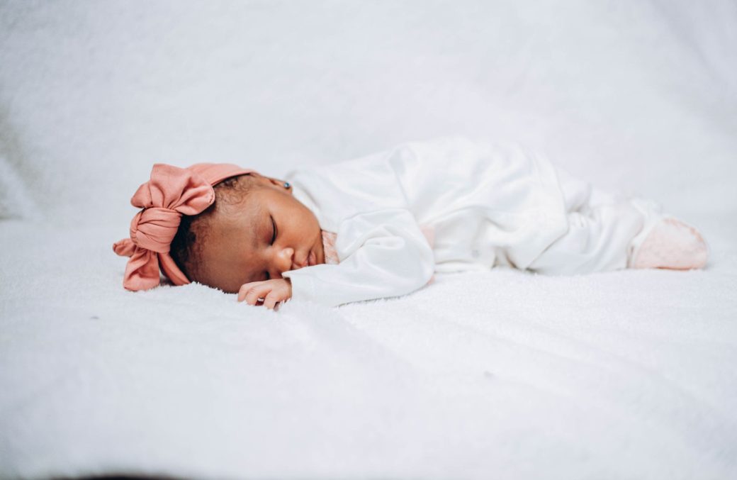 Adorable baby sleeping peacefully on white fabric, wearing a pink headband.