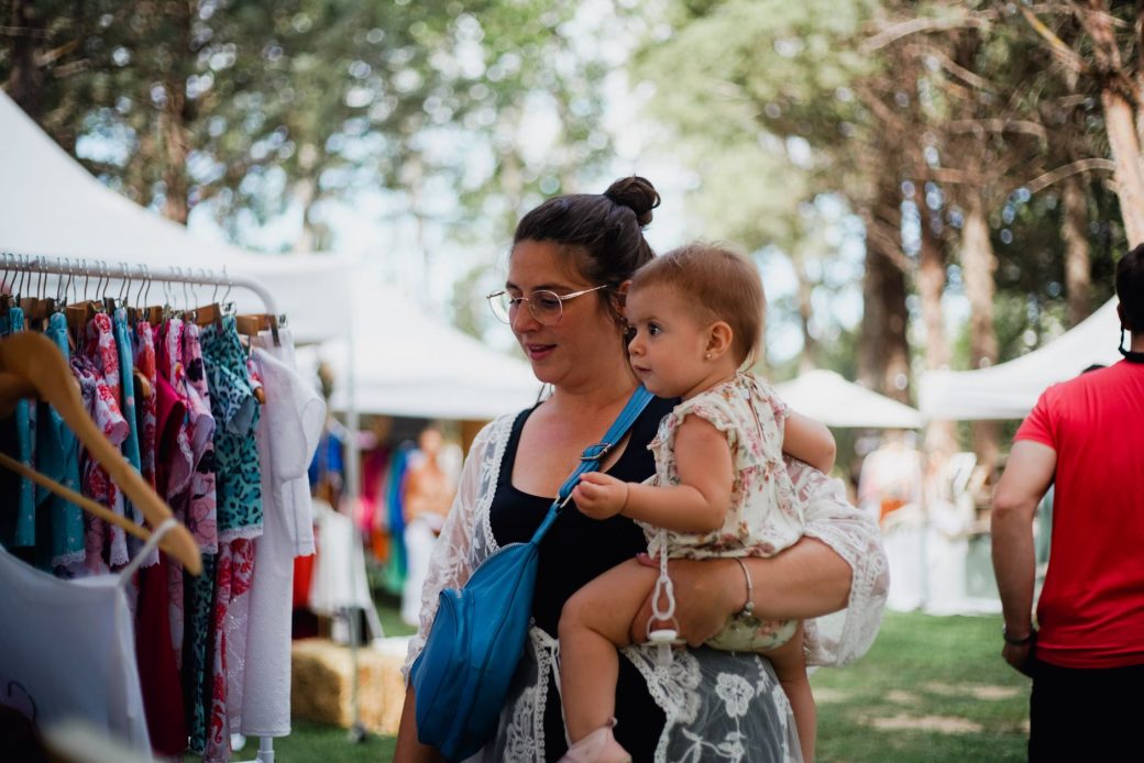 Mother with her baby shopping at an outdoor market surrounded by trees. Summer leisure activity.