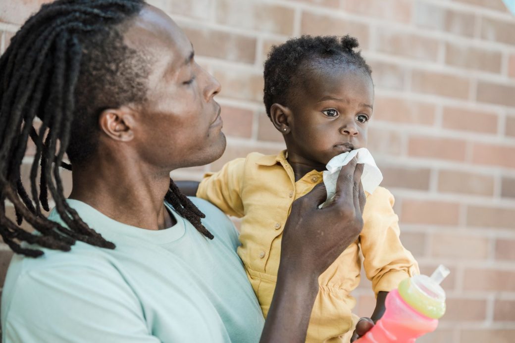 Man gently wiping young child's face while holding a bottle outdoors.