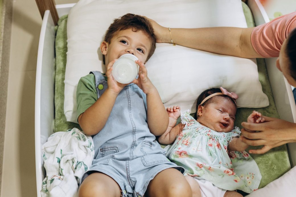 Young boy drinking milk beside his sleeping baby sister in a cozy bed setting.