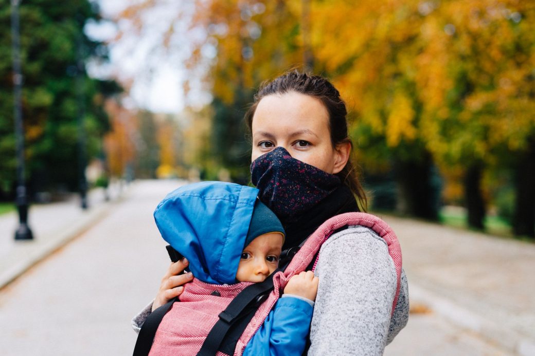 Woman wearing a mask carrying a baby in a carrier during a fall walk in the park.