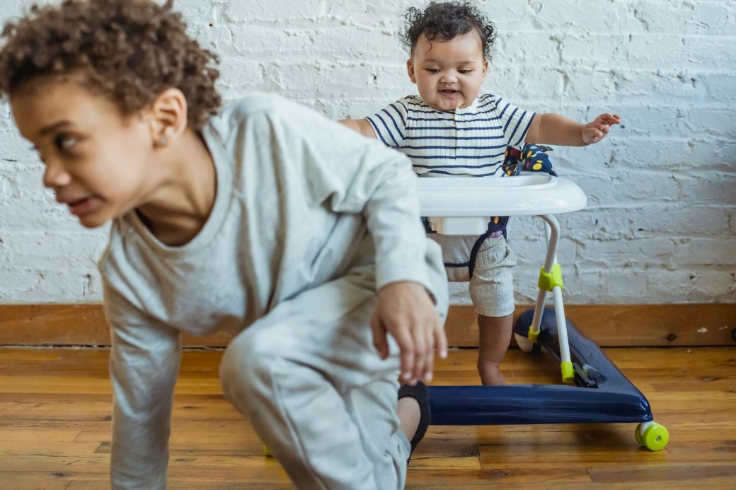 Playful African American boy in casual clothes near adorable little toddler in baby walker in light room near wall at home