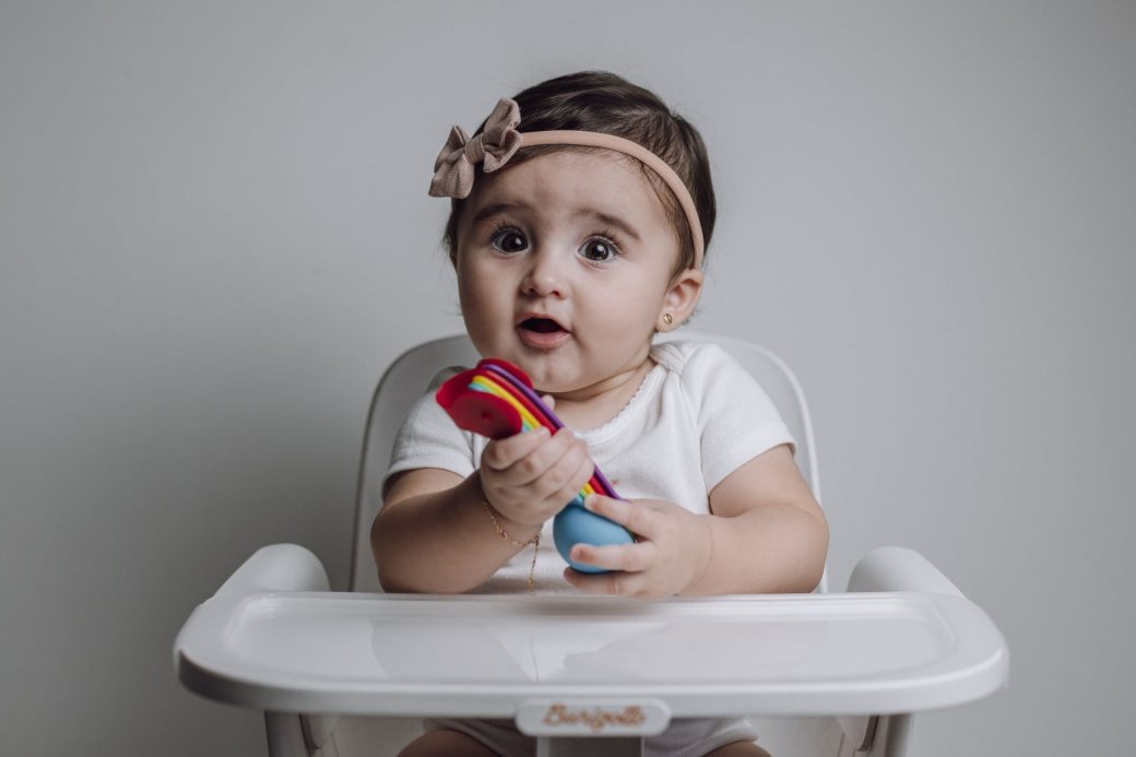 Charming baby girl with headband holding colorful toy while sitting in a high chair, studio setting.