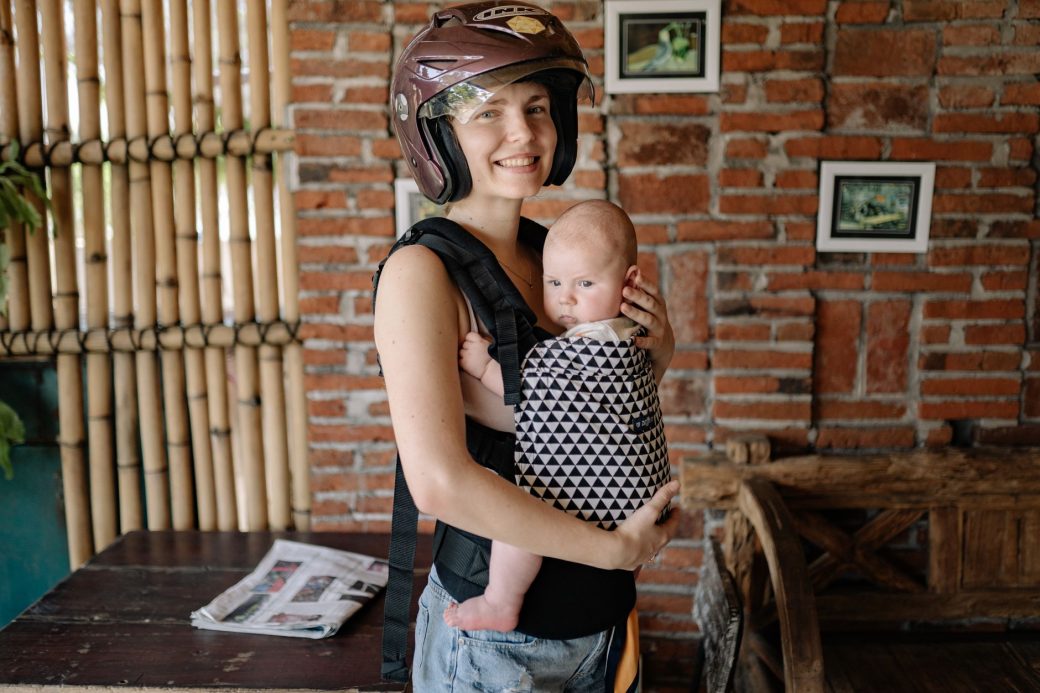 A smiling mother holding her baby in a carrier, both indoors with a helmet on.