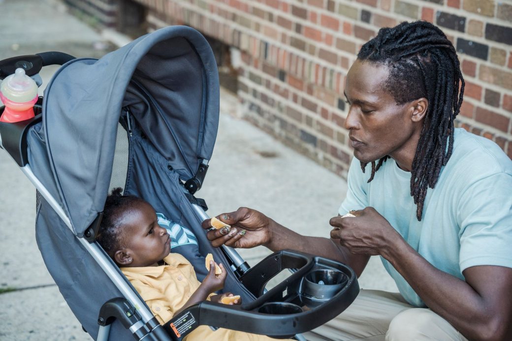 A father lovingly feeds his baby in a stroller outside against a brick wall.