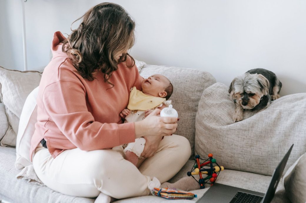 Side view of faceless female in casual clothes sitting on sofa with puppy and caressing adorable little baby while using laptop