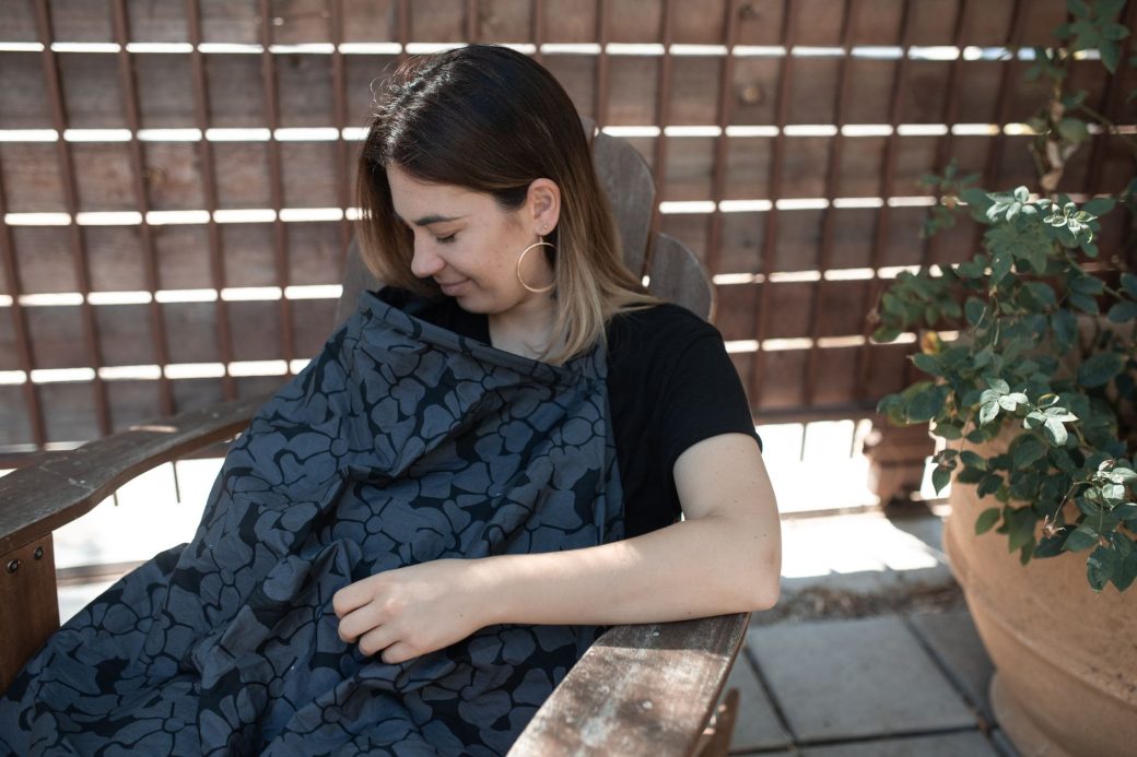 Mother sitting outdoors comfortably breastfeeding her baby under a stylish nursing cover.