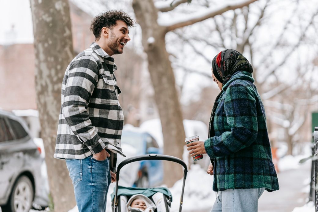 Side view of cheerful young ethnic parents standing near baby in stroller while spending winter day on snowy city street