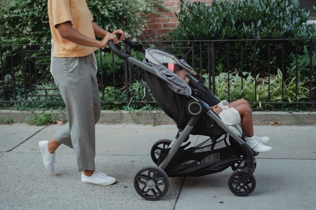 Crop anonymous ethnic female in casual clothes strolling on paved street of city with little girl in carriage