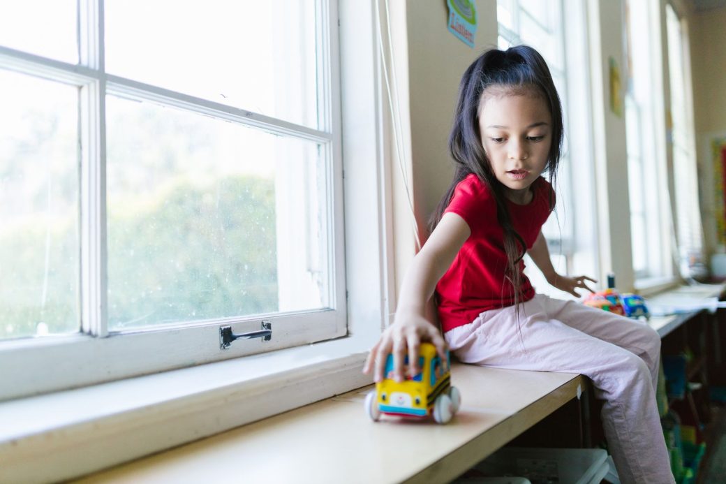 A young girl in a red shirt plays with a toy beside a window in a sunny classroom setting.