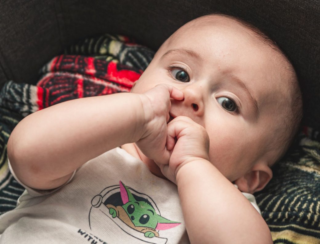 Cute baby in a onesie nibbling fingers, capturing innocence and playfulness.