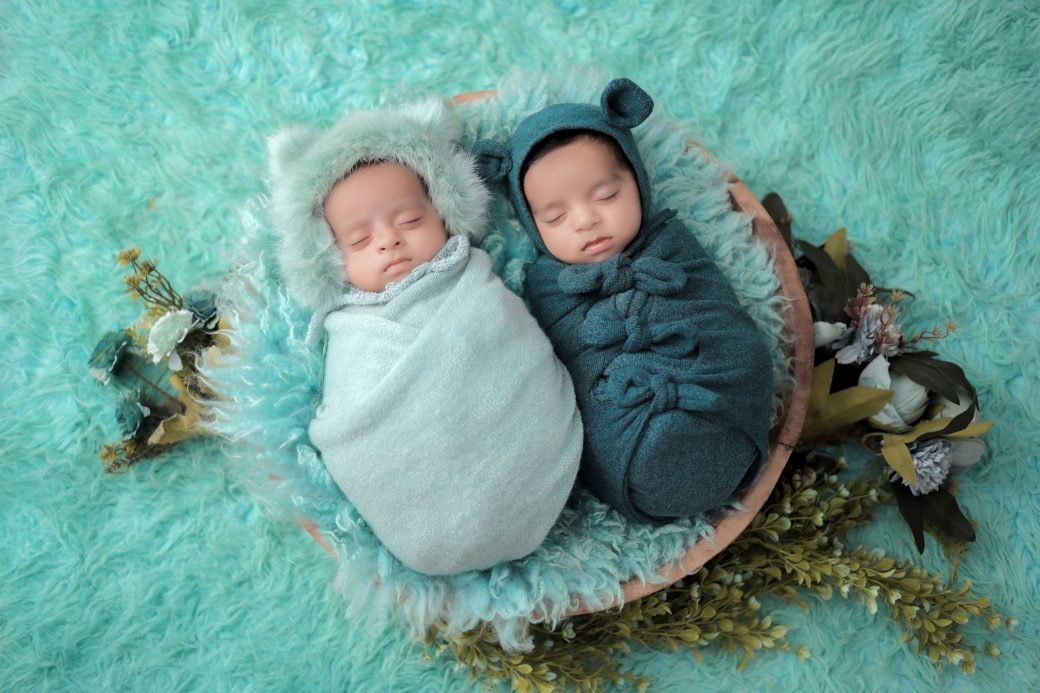 Two serene newborns wrapped in soft swaddles, sleeping peacefully on fluffy fabric with leafy decor.