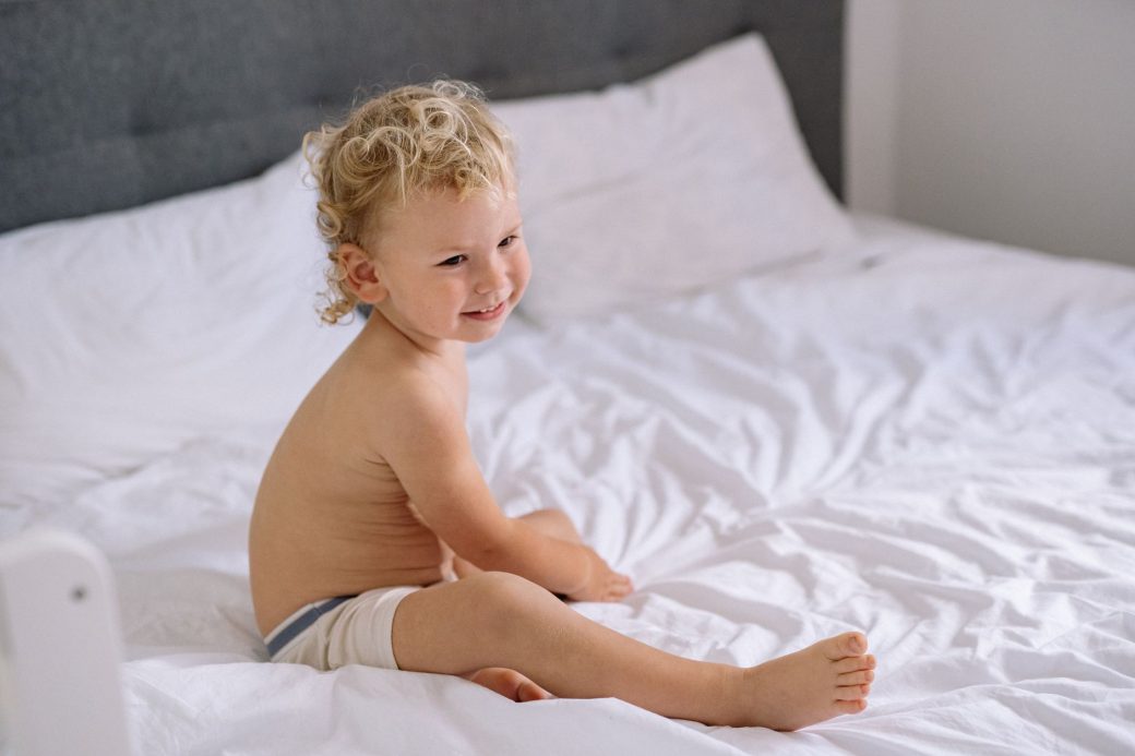 A smiling toddler with curly hair sitting happily on a white bed indoors.