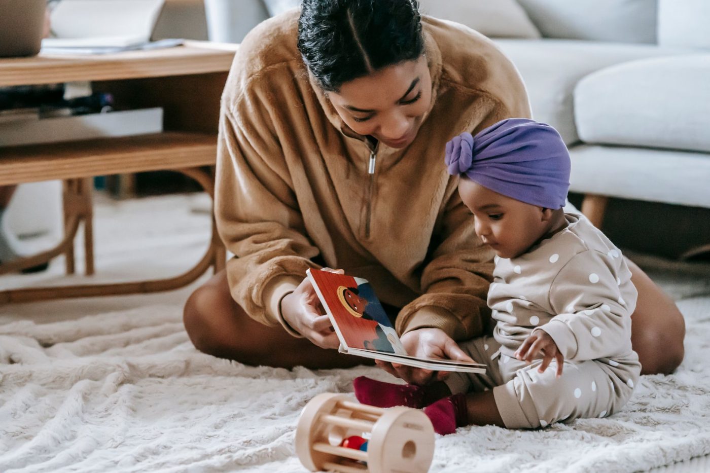 A mother showing a picture book to her young daughter while sitting on a rug at home.
